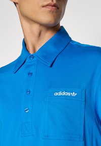 Mann som har på seg en klar blå Adidas polo med knapper og en logo-brodert brystlomme, vist fra nakken til midttorso.
