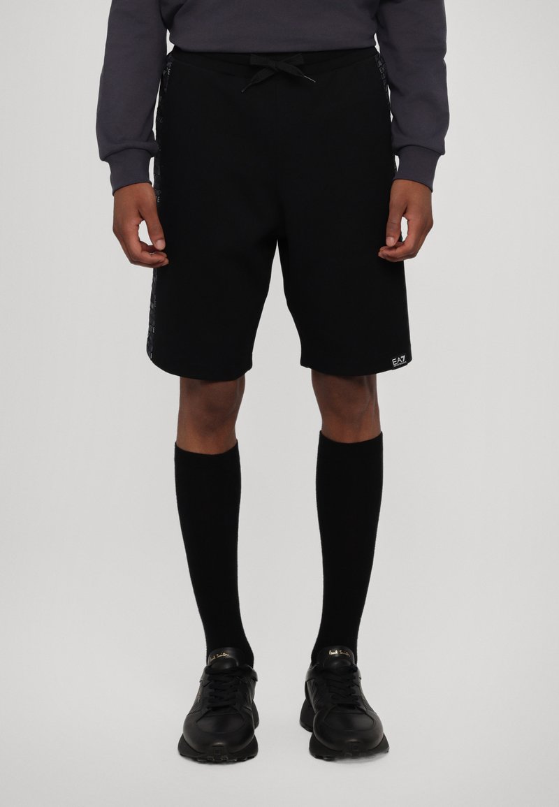 Shorts sportivi neri realizzati in tessuto morbido, con vita regolabile tramite coulisse e grafiche stampate sui lati. Abbinati a calze nere al ginocchio.