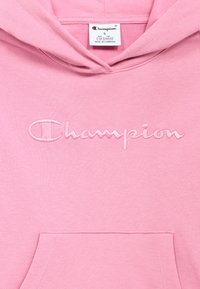 Ροζ φούτερ με κουκούλα, με κεντημένο λογότυπο "Champion" σε ανοιχτό ροζ χρώμα. Το υλικό είναι απαλό, με τσέπη καγκουρό στο μπροστινό μέρος.