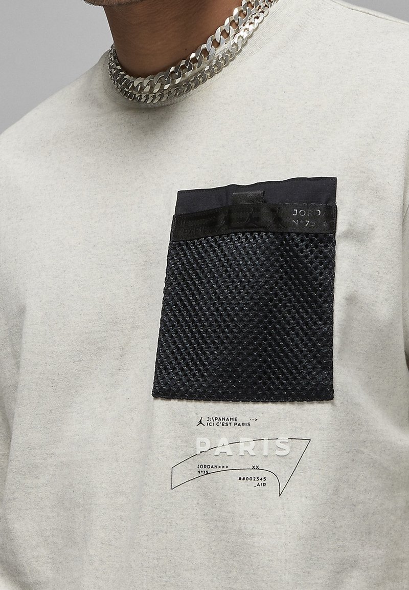 Camiseta de color gris claro con un bolsillo de malla negra texturizada, detalles serigrafiados en blanco y un collar de eslabones.