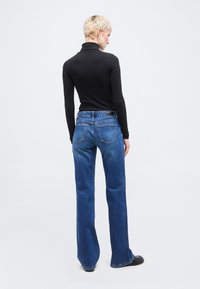 Ά τομο με κοντά ξανθά μαλλιά που φοράει μαύρο, μακρυμάνικο ζιβάγκο και μπλε τζιν με ίσια γραμμή, στέκεται με την πλάτη προς το μέρος μας σε ένα απλό φόντο.