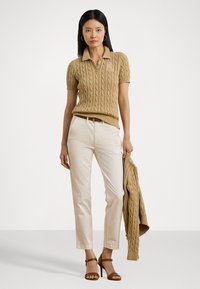 Beige kabelgebreide polo shirt, korte mouwen, geborduurd logo op de borst, gecombineerd met crème chino's en een bijpassende trui over één arm gedrapeerd.