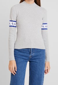 Maglietta grigia a maniche lunghe con righe e collo alto, decorata con strisce blu e loghi sulle maniche; abbinata a jeans blu.