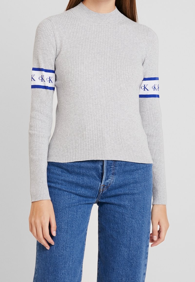 Graues, geripptes Langarmoberteil mit Stehkragen und blauen Streifen, die Logos auf den Ärmeln tragen; kombiniert mit blauen Jeans.