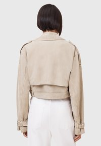 Beige Wildleder-Cropped-Jacke mit Stehkragen, Gurt-Detail in der Taille und Akzenten an den Ärmeln; glatte Textur und taillierte Passform.