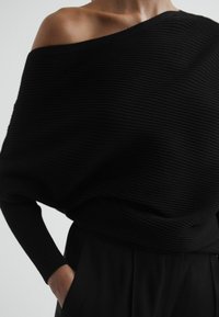 Zwarte geribbelde trui met een off-shoulder ontwerp en lange mouwen. Zachte textuur met een losse pasvorm, aangevuld met een high-waisted zwarte onderkant.