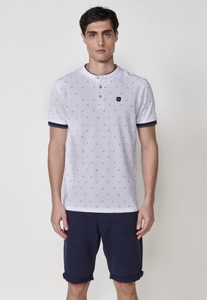 Polo bianco con piccolo motivo a triangoli neri, dotato di colletto con bottoni e rifiniture blu navy. Indossato con pantaloni corti blu navy, arrotolati all'orlo.