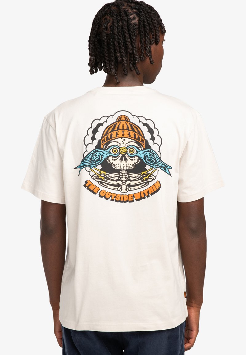 Camiseta de algodón crema con un gráfico colorido de un cráneo con gafas y un gorro, flanqueado por pájaros azules. El texto dice "EL EXTERIOR INTERIOR".