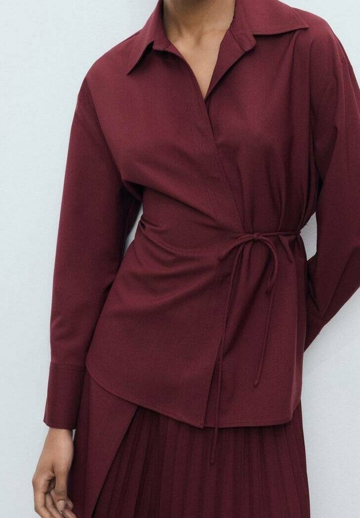 Femme portant un chemisier portefeuille à manches longues bordeaux avec col et ceinture à nouer, assorti à une jupe plissée de la même couleur.
