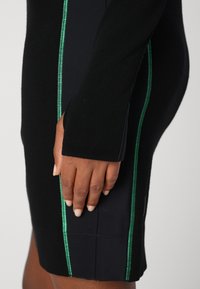 Schwarzes Kleid mit langen Ärmeln, ausgestattet mit einem grünen Seitenstich-Detail und einem strukturierten schwarzen Stoff. Glatte Oberfläche, taillierte Passform.