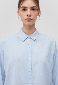 Hellblaues Hemd mit Knopfleiste, strukturiertem Kragen und kleinen blauen Punktmustern. Aus einem strukturierten, leichten Material gefertigt. Weiße Knöpfe sind vorhanden.