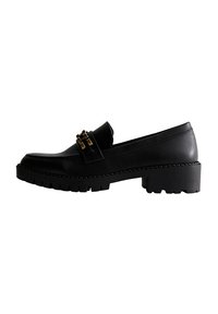 Mocassino in pelle nera con suola spessa e sagomata, caratterizzato da un accessorio decorativo in metallo dorato sulla fascia. Finitura liscia, punta rotonda.