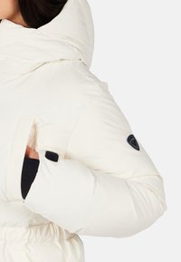 Nærbillede af en person iført en hvid polstret jakke med høj krave, sort indvendig lomme og en sort logo-patch på ærmet.
