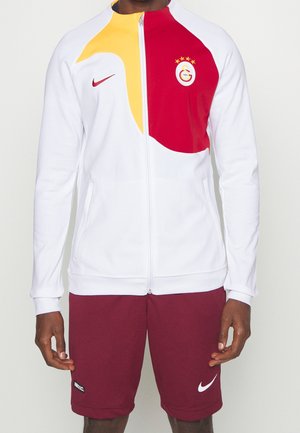 Biała kurtka na zamek z żółto-czerwonym wzorem, logo Galatasaray na piersi, w połączeniu z bordowymi szortami Nike z białymi logo Swoosh.
