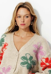 Cardigan en tricot beige avec des motifs floraux rouges, verts et roses. Présente un col en V et deux boutons à l'avant. Texture douce.