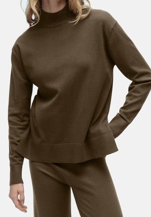 Femme portant un haut en maille à manches longues marron et un pantalon assorti, main sur la hanche, debout devant un fond clair uni.