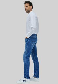 Jeans de mezclilla azul de corte slim, con cinco bolsillos y lavado desgastado. Combinados con una camisa blanca de manga larga con botones y zapatillas blancas.