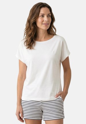 Vrouw met een effen wit T-shirt met korte mouwen en marineblauw-wit gestreepte korte broek, staand met één hand in de zak, kijkend naar rechts.