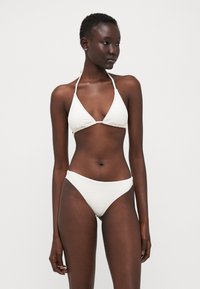 Bijela bikini kombinacija koja sadrži teksturiranu trokutaastu majicu i klasične donje dijelove niskog struka, s tankim trakama i minimalnim pokrivanjem u jednostavnom dizajnu.