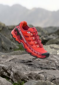 Παπούτσι για τρέξιμο σε μονοπάτια σε έντονο κόκκινο-πορτοκαλί πλέγμα και καουτσούκ. Διαθέτει υφασμένο πέλμα και εμφανές σύστημα κορδονιών. Ελαφρύς σχεδιασμός για απόδοση.