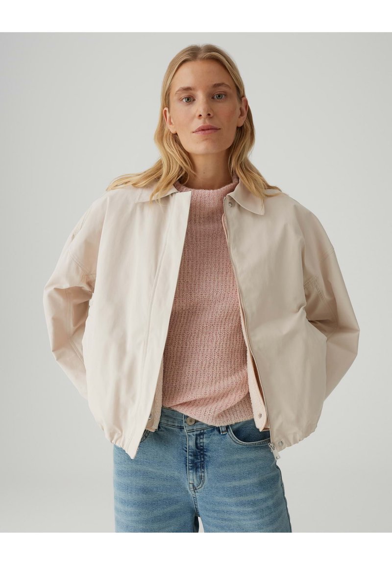 Lichtroze, met rits gesloten bomberjack met een lichte glans, voorzien van een kraag en elastische manchetten. Draagt over een gestructureerde roze trui en blauwe jeans.