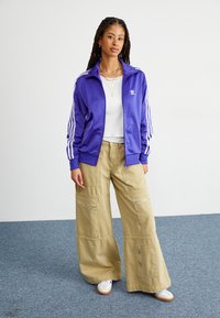 Giacca a track in satin viola con strisce bianche e logo, abbinata a pantaloni cargo beige a gamba larga con zip e tasche. Sneakers bianche completano l'outfit.