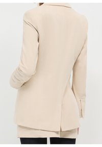 Beżowa marynarka ze sztruksu z klapą z wycięciem, długimi rękawami, rozcięciem z tyłu i fakturowaną tkaniną. Charakteryzuje się dopasowanym krojem i czystymi liniami.
