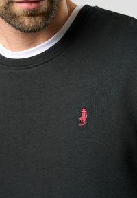 Zwarte sweatshirt met een geribde halslijn en een witte binnenlaag, voorzien van een klein roze geborduurd logo op de linkerborst.