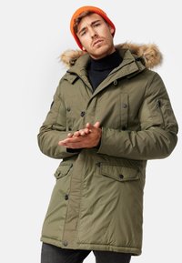 Parka verde oliva con borde de capucha de pelo sintético, cierres de cremallera y broches a presión, múltiples bolsillos frontales y detalles de hardware negro. Tejido texturizado.
