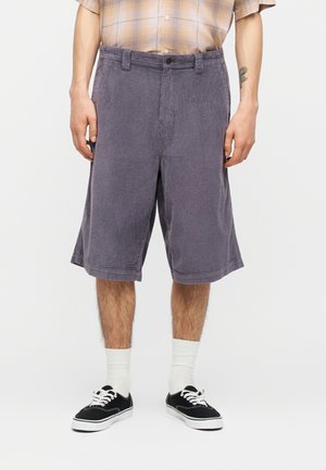 Bărbat purtând pantaloni scurți gri din catifea cu talie medie și lejeri, șosete albe, adidași negri și o cămașă bej în carouri băgată în pantaloni.