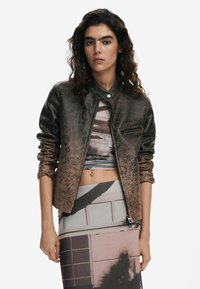 Veste brune texturée avec un col mandarin, poche zippée et manches retroussées. Le design présente un motif abstrait.