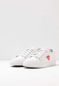 Vita kanvas sneakers med rund tå, platt sula, snörning och en röd hjärt-grafik på sidan. Texturerat tyg och minimalistisk design.