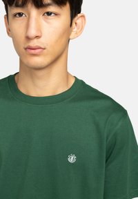Zelené bavlněné tričko s kulatým výstřihem a krátkými rukávy, s malým bílým logem na levé straně hrudi. Hladká textura.