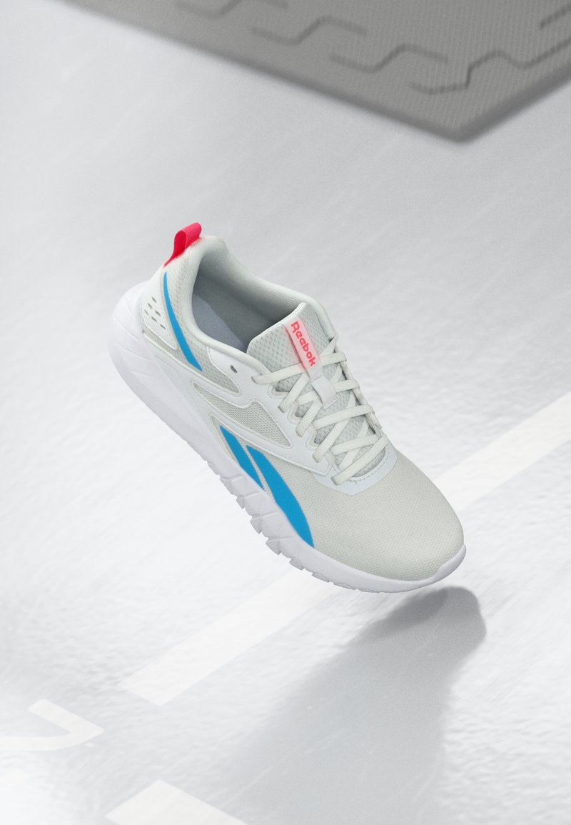Sneaker Reebok bianco e blu con un pull tab rosso che galleggia sopra un pavimento lucido con un tappetino di gomma grigio nelle vicinanze.