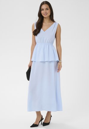 Femme aux longs cheveux noirs portant une robe bleu clair sans manches avec basque, talons noirs pointus, tenant une pochette noire tressée, souriante, fond uni.