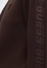 Gros plan d'un vêtement marron avec une fermeture éclair, un logo triangulaire et un ruban texturé portant le nom de la marque "GUESS" sur la manche.