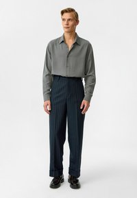 Grijs overhemd met knopen en een soepele textuur, gecombineerd met marineblauwe gestreepte broek die een losse pasvorm heeft en omgeslagen manchetten. Zwarte schoenen maken de look compleet.