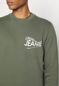 Sudadera verde oliva con cuello redondo. Presenta el texto blanco "CALVIN KLEIN JEANS" con letras curvas y un tejido suave y texturizado.