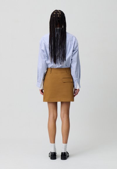 Gonna in cotone marrone con un taschino sul retro, abbinata a una camicia a quadri blu. Il design è dritto e ha una lunghezza sopra il ginocchio.