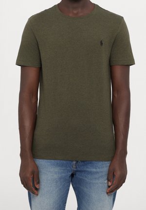 Mężczyzna ubrany w oliwkowo zielony t-shirt z krótkim rękawem i okrągłym dekoltem oraz jasnoniebieskie jeansy na białym tle.