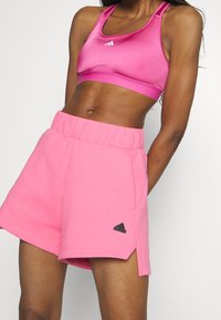 Vaaleanpunainen urheilurintaliivit ja vastaavat shortsit pehmeästä kankaasta. Shortseissa on joustava vyötärönauha ja sivuhalkio, jossa on pieni musta logokoriste.