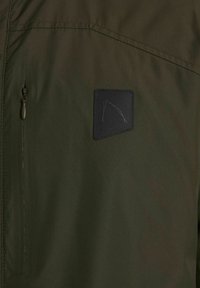 Chaqueta verde oscura con un tejido texturizado, bolsillo con cremallera y un parche negro con un logotipo. Diseño sencillo y funcional sin patrones visibles.