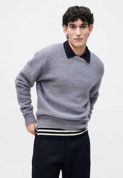 Maglione grigio in misto lana con collo scuro, caratterizzato da un orlo a coste con strisce bianche e nere, indossato sopra pantaloni neri a righe blu.