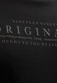 Svart stoff med hevet tekst som lyder "Nineteen ninety Original 1990 Brought to you by Jac" i en teksturert, preget stil.