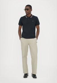Polo cu mânecă scurtă, de culoare bleumarin, cu dungi bej la guler, asortat cu pantaloni bej deschis și pantofi negri. Modelul poartă ochelari de soare.