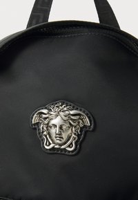 Fekete nylon hátizsák, amelyen egy kiemelkedő ezüst Medúza fej embléma található. Simább textúra, cipzáras záródással és állítható pántokkal.