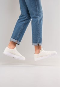 Weiße Mesh-Sneaker mit einem niedrig-profiligen Design, ausgestattet mit einem strukturierten Obermaterial, runder Zehenpartie und einer weißen Gummisohle, getragen mit hochgekrempelten blauen Jeans.