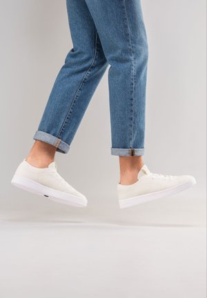 Białe siatkowe sneakersy o niskim profilu, z teksturowaną cholewką, okrągłym noskiem i białą gumową podeszwą, noszone z podwiniętymi niebieskimi dżinsami.
