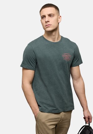 Jeune homme portant un T-shirt graphique vert foncé et un pantalon beige, tenant un sac noir dans sa main droite, regardant sur le côté.