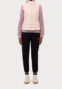 Person som bär en ljusrosa quiltad väst över en mauve långärmad skjorta, svarta joggers med logotyp och krämfärgade sneakers med gummisulor, stående rakt.